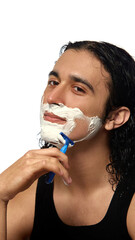 Young man with curly wet hair shaving face with blue razor, thick foam covering skin against white...
