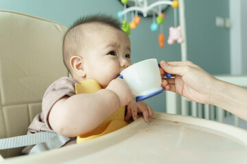 Asian Baby drinking from cup in high chair, assisted by hand, learning to drink, feeding time