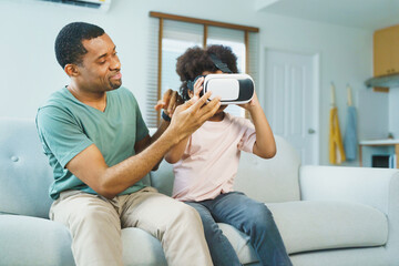 Black African American Father assisting son with virtual reality headset on a living room sofa, engaged in a playful learning activity