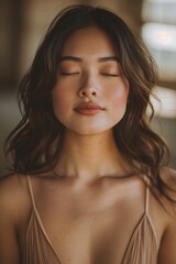 Fototapeta premium Serene young Asian woman with her eyes closed, wearing a beige camisole. Soft lighting and warm tones create a peaceful mood.
