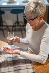 Senior woman sitting at a kitchen table, holding a glass of water and looking at medication in her hand. She appears thoughtful and focused on her health and wellness