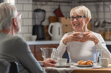 Senior couple enjoying breakfast together in a cozy kitchen. The elderly woman, smiling, holds a cup of coffee while chatting with her partner, creating a warm and relaxed atmosphere.