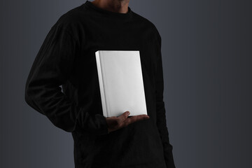 A person wearing a black long-sleeve shirt, holding an A5-sized book mockup in front of their torso. Isolated blank mockup surface to place your design. 