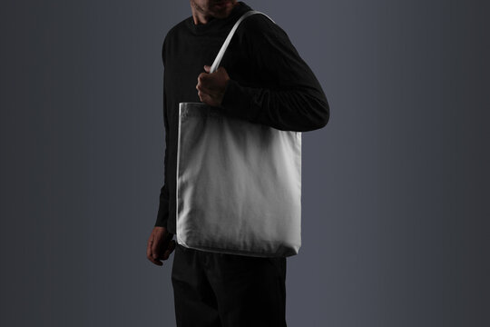 A tote bag mockup, carried by a man dressed in black attire, including a long-sleeved shirt, and pants. Isolated blank mockup surface to place your design. 