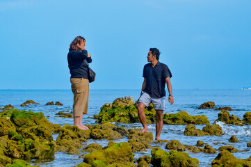 A man and a woman are standing barefoot on a mossy, rocky shoreline, engaged in conversation while...