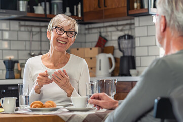 Senior couple enjoying breakfast together in a cozy kitchen. The elderly woman, smiling, holds a cup of coffee while chatting with her partner, creating a warm and relaxed atmosphere.