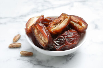 Many tasty dried dates with seeds in bowl on white marble table, closeup