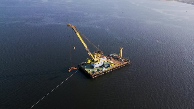 A floating crane barge with a large yellow crane operates on calm waters, lifting an object with its hook. The industrial vessel is surrounded by an open water expanse with a distant shoreline
