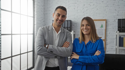 Smiling man and woman standing confidently in an office environment with arms crossed showcasing a...