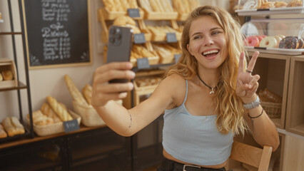 Young woman taking a selfie inside a bakery, smiling and posing with a peace sign, surrounded by fresh bread and pastries on display