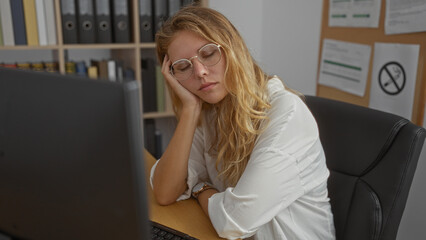 Attractive blonde woman sleeping at her desk in an office room with shelves filled with binders in...