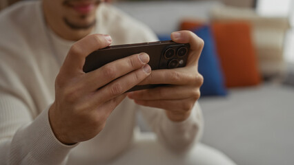 Young man holding smartphone indoors with hands visible in modern living room setting, suggesting relaxed home environment and digital interaction focus