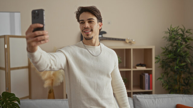 Young hispanic man in cozy apartment living room taking selfie with smartphone capturing casual moment indoors