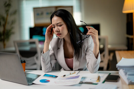 Professional businesswoman experiencing work stress, pressing temples while seated at laptop in corporate workspace