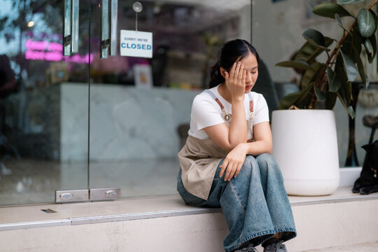 Young asian waitress sitting with head in hand in front of her closed store, showing concern and financial loss - Powered by Adobe