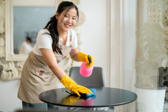 Young woman wearing apron and gloves cleaning table surface with disinfectant spray and blue cloth, ensuring hygiene in restaurant
