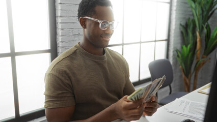 Young man with glasses sitting indoors at an office desk counting american money with a smile,...