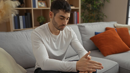 Young man with contemplative expression sits in a cozy living room, surrounded by plush pillows and...