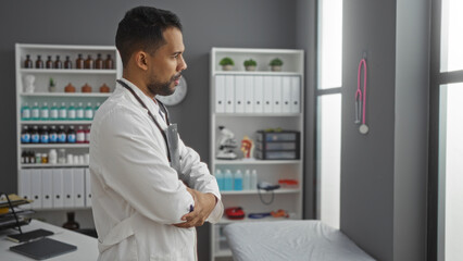 Fototapeta premium Handsome young man stands with arms crossed in a modern clinic setting, exuding professionalism and confidence within a healthcare environment.