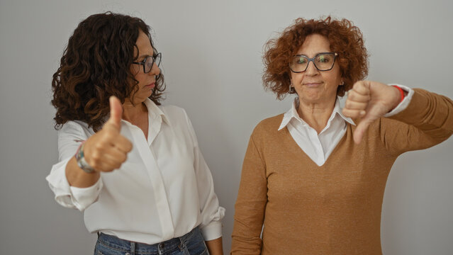 Women expressing different opinions with thumbs up and down against a white wall, showcasing diverse reactions among two middle-aged friends on an isolated background.