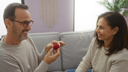 Middle-aged hispanic couple sitting together indoors, man holding ring box proposing to smiling woman on sofa in cozy home living room.