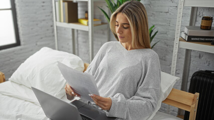 Woman reading document on bed in modern bedroom using laptop showing a relaxed and focused atmosphere in a bright indoor setting.