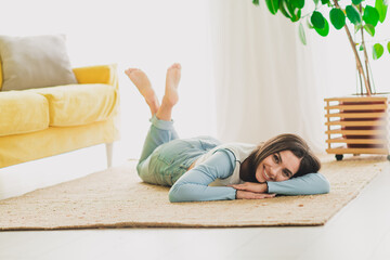 Smiling young woman lying on a cozy rug in a bright and modern living room, enjoying a moment of...