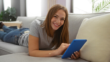 Woman smiling indoors while using a tablet in a cozy living room, showcasing a relaxed and modern...