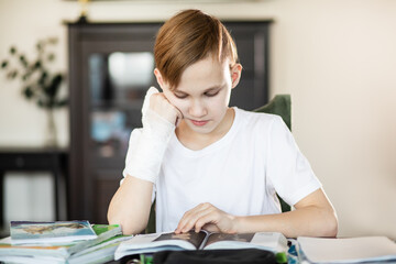 Kid teenager boy pupil at home making homework with broken finger. Child hand with a cast.