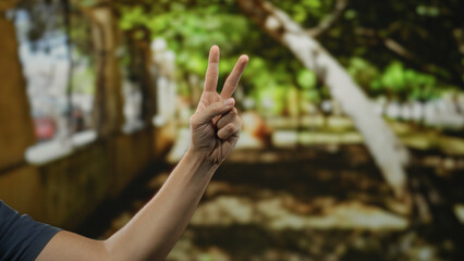 Man showing peace gesture with hand outdoors against blurred park background in sunny warm weather capturing calm vibrant nature atmosphere.