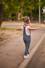 The girl walks along the sidewalk, keeping her balance, arms outstretched.
