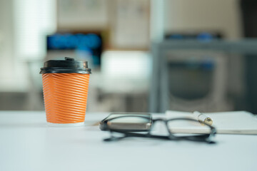 Disposable orange coffee cup standing on white desk with eyeglasses, notepad and pen in blurred office background representing break time or morning routine