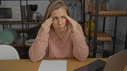Woman stressed in office wearing sweater holds head while sitting at desk with laptop, expressing...