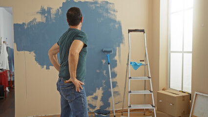 Middle-aged hispanic man in new home living room looking at a half-painted wall with a roller and...