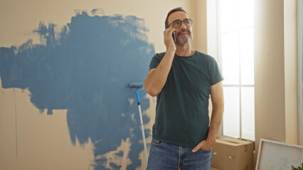 Mature man talking on phone standing in a newly painted room of a home with paint roller in...