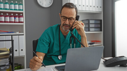 Hispanic middle-aged man in a clinic wearing glasses and green scrubs is working on a laptop while talking on a phone, showcasing a professional and healthcare environment.