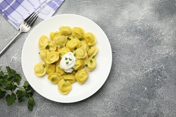 Tasty dumplings with green onion, sour cream and fork on grey textured table, flat lay. Space for text