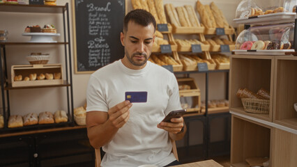 Young hispanic man with beard in bakery holding credit card and mobile phone surrounded by bread pastries in indoor cafe setting