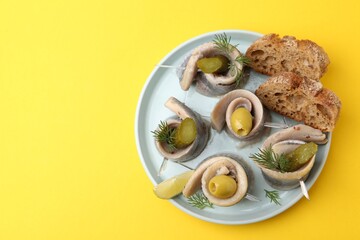 Delicious herring rolls with olives, pickles, dill and bread pieces on yellow table, top view....