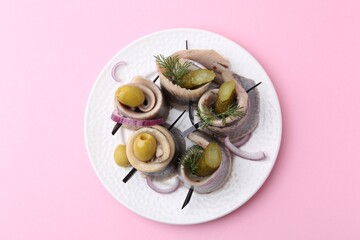 Delicious herring rolls with olives, pickles, dill and onion on pink table, top view