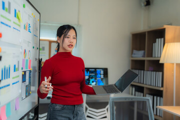 Young professional woman holding a laptop while explaining charts and graphs on a whiteboard during a dynamic presentation in a modern office environment