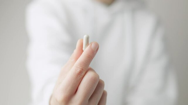 A woman shows a vitamin D capsule pill, with her right hand to the camera, she wears a white sweatshirt on white background