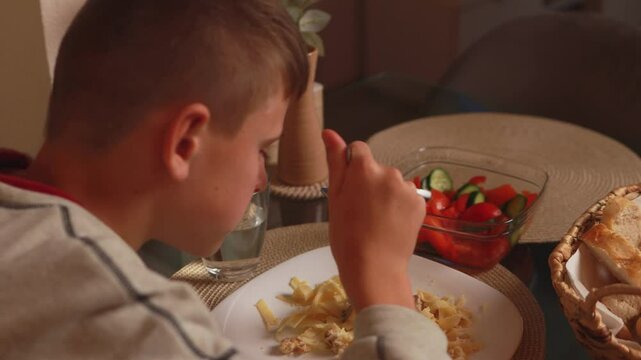Young boy savoring colorful pasta, eagerly consuming homemade meal at kitchen table with pure culinary delight