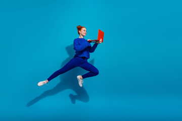 Young woman with red hair jumping energetically on blue background holding a laptop, radiating...