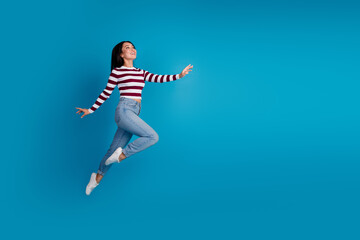 Obraz premium Smiling young woman leaping with joy against a solid blue background wearing trendy casual striped shirt and jeans