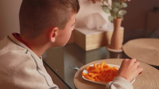 Young boy munching corn puffs, sitting casually at kitchen table, experiencing relaxed snacking moment