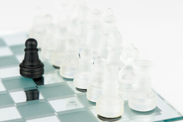 A lone black pawn opposes a formation of white chess pieces on a glass chessboard. The mirrored...