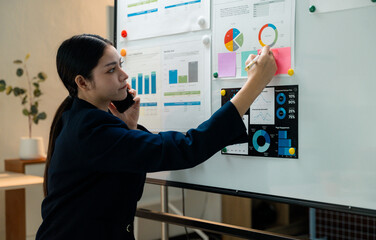 Businesswoman writing on whiteboard and analyzing financial charts while talking on smartphone