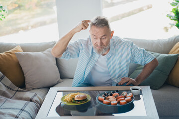 Elderly man pondering a dining choice between sushi and a burger in a cozy living room setting under daylight