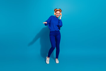Stylish young woman with red hair smiling and enjoying music on headphones against a vibrant blue...